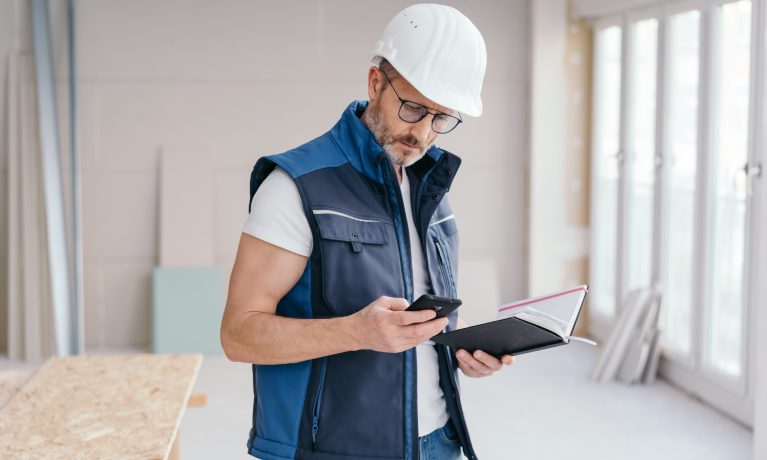 Builder checking a message on his mobile phone or making a call as he stands holding a journal on site in a new build house